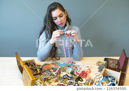 young brunette sitting at the table with lots of jewelry, choosing and trying different jewelry young brunette sitting at the table with lots of jewelry, choosing and trying different jewelry 126354500