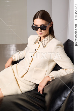 Stylish female fashion model sits on sofa confidently in a trendy beige outfit and sunglasses indoors Stylish female fashion model sits on sofa confidently in a trendy beige outfit and sunglasses indoors 126354635