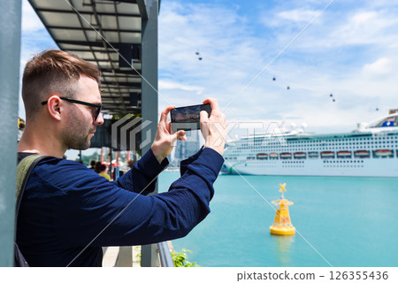Russian tourist man taking photo of a cruise ship at Sentosa Island in Singapore on a sunny day with cable cars in the background. Russian tourist man taking photo of a cruise ship at Sentosa Island in Singapore on a sunny day with cable cars in the background. 126355436
