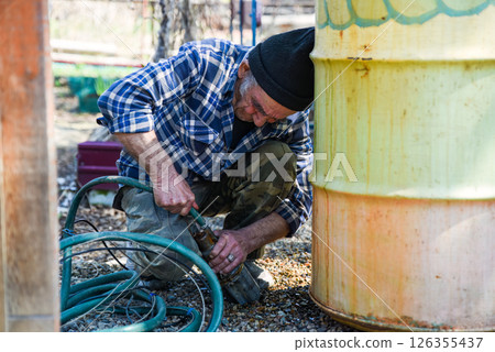 Elderly Russian man sitting near a barrel pulling a drainage hose to the side with visible tension and effort in a quiet rural setting. Elderly Russian man sitting near a barrel pulling a drainage hose to the side with visible tension and effort in a quiet rural setting. 126355437