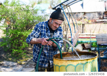 Elderly Russian man pulling a drainage pump hose from a barrel with visible effort, doing physical work in a village backyard. Elderly Russian man pulling a drainage pump hose from a barrel with visible effort, doing physical work in a village backyard. 126355438