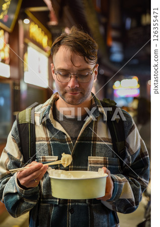 Russian tourist holding a crab and pork dumpling with chopsticks, looking at it before eating, on Yuyuan Old Street in Shanghai, wearing a plaid shirt Russian tourist holding a crab and pork dumpling with chopsticks, looking at it before eating, on Yuyuan Old Street in Shanghai, wearing a plaid shirt 126355471