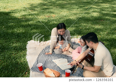 Parents read a book to their young children while relaxing on a blanket outdoors Parents read a book to their young children while relaxing on a blanket outdoors 126356101