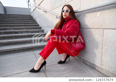 High fashion photo of a young brunette woman in red glossy down jacket, and red pants. Urban street fashion. High fashion photo of a young brunette woman in red glossy down jacket, and red pants. Urban street fashion. 126357674