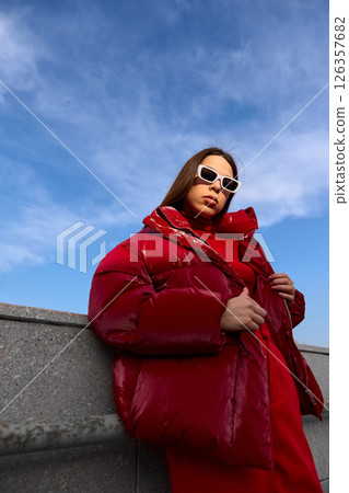 High fashion photo of a young brunette woman in red glossy down jacket, and red pants. Urban street fashion. High fashion photo of a young brunette woman in red glossy down jacket, and red pants. Urban street fashion. 126357682