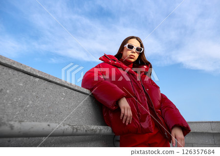 High fashion photo of a young brunette woman in red glossy down jacket, and red pants. Urban street fashion. High fashion photo of a young brunette woman in red glossy down jacket, and red pants. Urban street fashion. 126357684