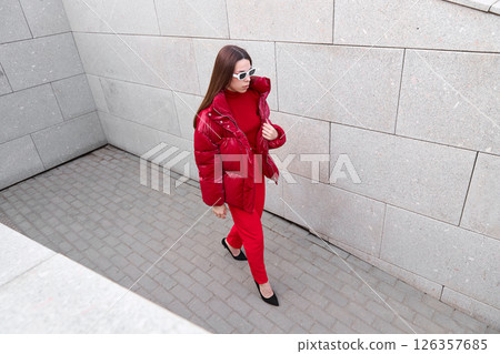 High fashion photo of a young brunette woman in red glossy down jacket, and red pants. Urban street fashion. High fashion photo of a young brunette woman in red glossy down jacket, and red pants. Urban street fashion. 126357685