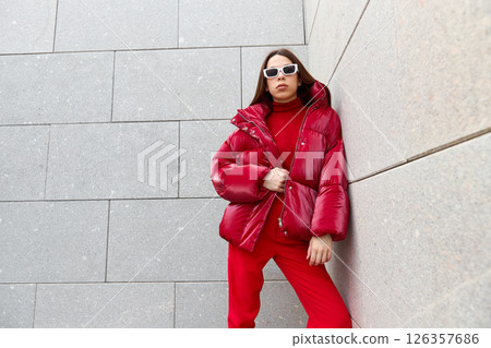 High fashion photo of a young brunette woman in red glossy down jacket, and red pants. Urban street fashion. High fashion photo of a young brunette woman in red glossy down jacket, and red pants. Urban street fashion. 126357686