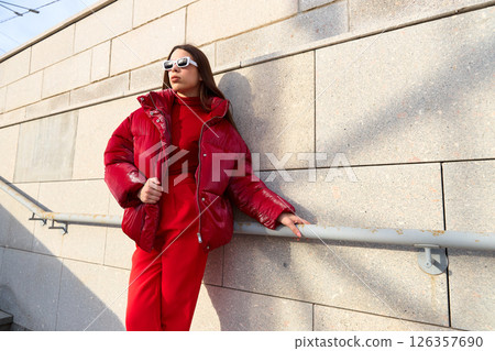 High fashion photo of a young brunette woman in red glossy down jacket, and red pants. Urban street fashion. 126357690