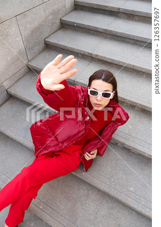High fashion photo of a young brunette woman in red glossy down jacket, and red pants. Urban street fashion. High fashion photo of a young brunette woman in red glossy down jacket, and red pants. Urban street fashion. 126357697