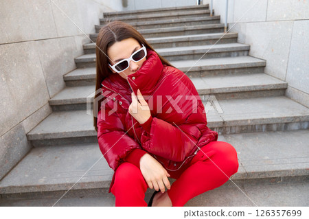 High fashion photo of a young brunette woman in red glossy down jacket, and red pants. Urban street fashion. High fashion photo of a young brunette woman in red glossy down jacket, and red pants. Urban street fashion. 126357699