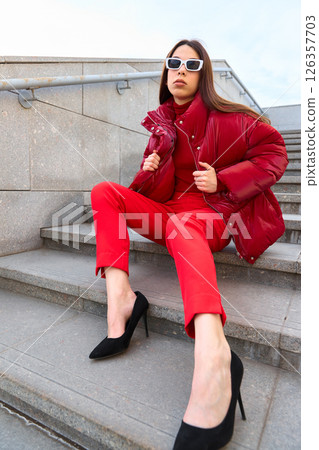 High fashion photo of a young brunette woman in red glossy down jacket, and red pants. Urban street fashion. High fashion photo of a young brunette woman in red glossy down jacket, and red pants. Urban street fashion. 126357703