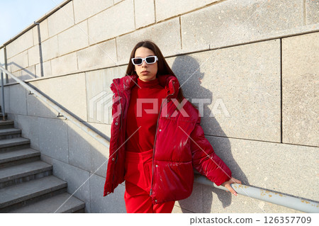 High fashion photo of a young brunette woman in red glossy down jacket, and red pants. Urban street fashion. 126357709