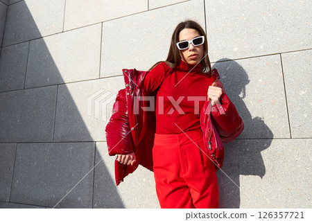 High fashion photo of a young brunette woman in red glossy down jacket, and red pants. Urban street fashion. High fashion photo of a young brunette woman in red glossy down jacket, and red pants. Urban street fashion. 126357721