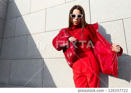 High fashion photo of a young brunette woman in red glossy down jacket, and red pants. Urban street fashion. High fashion photo of a young brunette woman in red glossy down jacket, and red pants. Urban street fashion. 126357722