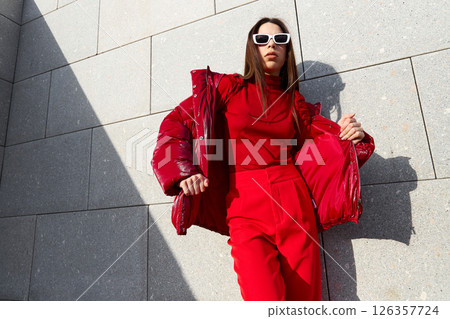 High fashion photo of a young brunette woman in red glossy down jacket, and red pants. Urban street fashion. High fashion photo of a young brunette woman in red glossy down jacket, and red pants. Urban street fashion. 126357724