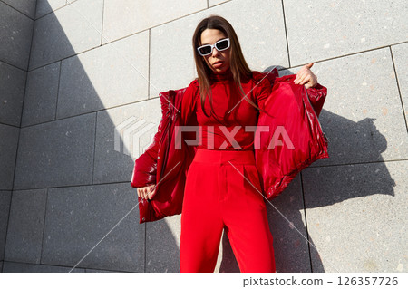 High fashion photo of a young brunette woman in red glossy down jacket, and red pants. Urban street fashion. 126357726