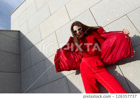 High fashion photo of a young brunette woman in red glossy down jacket, and red pants. Urban street fashion. 126357737