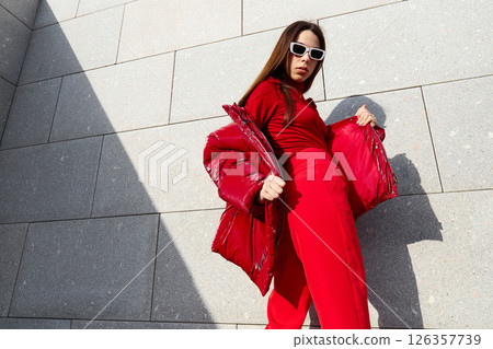 High fashion photo of a young brunette woman in red glossy down jacket, and red pants. Urban street fashion. 126357739