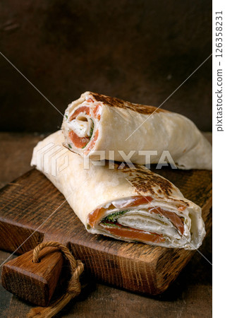 Delicious salmon lavash wrap with cream cheese fresh greens and vegetables served on rustic wooden cutting board. Homemade appetizer with smoked salmon. 126358231