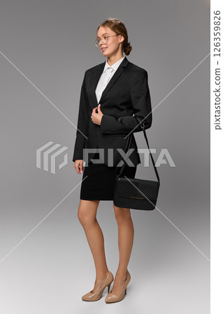 Elegant woman in business outfit holding black handbag and smiling slightly against grey background. Elegant woman in business outfit holding black handbag and smiling slightly against grey background. 126359826