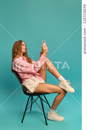 Young woman sitting sideways on chair smiling at phone screen in pink shirt and white skirt on teal background. Young woman sitting sideways on chair smiling at phone screen in pink shirt and white skirt on teal background. 126359836