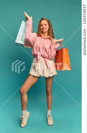 Happy young woman in pink shirt proudly showing shopping bags and smiling confidently against teal backdrop. Happy young woman in pink shirt proudly showing shopping bags and smiling confidently against teal backdrop. 126359837