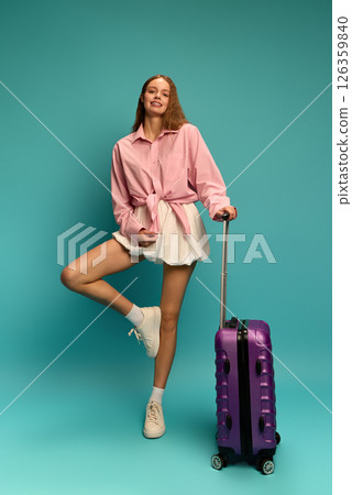 Cheerful young woman posing with purple suitcase, wearing pink shirt and white skirt, lifting leg playfully against teal background Cheerful young woman posing with purple suitcase, wearing pink shirt and white skirt, lifting leg playfully against teal background 126359840