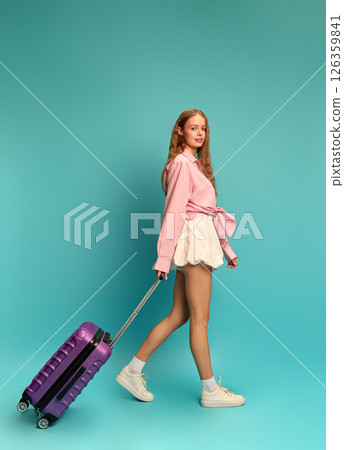 Young stylish woman in pink shirt and skirt walking with purple suitcase against teal backdrop. Vacation. Young stylish woman in pink shirt and skirt walking with purple suitcase against teal backdrop. Vacation. 126359841