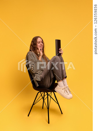 Woman sitting cross-legged on black chair holding tablet for video call smiling and waving against yellow background 126359856