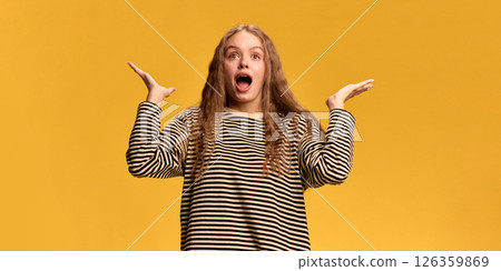 Young woman expressing surprise and shock with her mouth open and hands raised in gesture of disbelief against yellow studio background. 126359869