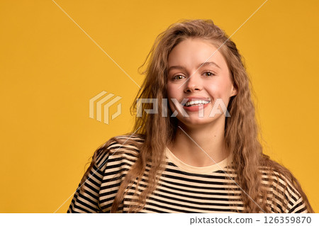 Close-up portrait of cheerful young woman with wavy hair, smiling, expressing happiness against yellow background. Close-up portrait of cheerful young woman with wavy hair, smiling, expressing happiness against yellow background. 126359870