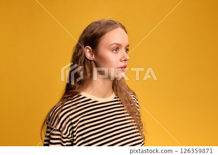 Portrait of young woman with long hair, in striped shirt looking sideways with calm expression against yellow background. 126359871