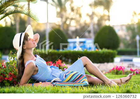 Young woman wearing light blue summer dress and yellow straw hat relaxing on green grass lawn in summer park. Girl in casual outfit resting outdoors enjoying free time in warm morning. Young woman wearing light blue summer dress and yellow straw hat relaxing on green grass lawn in summer park. Girl in casual outfit resting outdoors enjoying free time in warm morning. 126360732