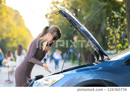 Young upset woman driver talking on mobile phone near a broken car with open hood waiting for help having trouble with her vehicle on a city street. 126360736