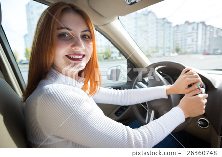 Young redhead woman driver behind a wheel driving a car smiling happily. 126360748