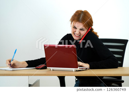 Young office worker woman talking on a cell phone sitting behind working desk with laptop computer and notebook. 126360749