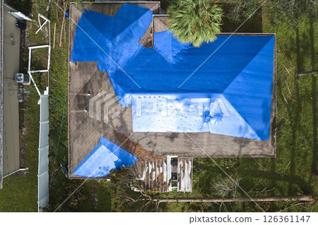 Top view of leaking house roof covered with protective tarp sheets against rain water leaks until replacement of asphalt shingles. Damage of building rooftop as aftermath of hurricane Ian in Florida Top view of leaking house roof covered with protective tarp sheets against rain water leaks until replacement of asphalt shingles. Damage of building rooftop as aftermath of hurricane Ian in Florida 126361147