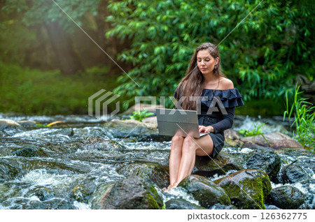 woman freelancer sitting on a rock in the river and using a laptop, travel or vacation work concept woman freelancer sitting on a rock in the river and using a laptop, travel or vacation work concept 126362775