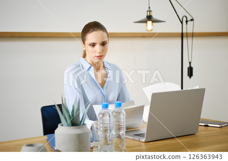 Focused woman reviewing documents at a modern desk setup with a laptop in a well-lit office 126363943