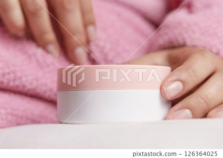 Woman in pink bathrobe puts white cream jar on white bedsheet closeup, cosmetic mockup Woman in pink bathrobe puts white cream jar on white bedsheet closeup, cosmetic mockup 126364025