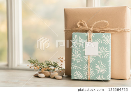 Two beautifully wrapped gifts with natural twine bows sit on wooden surface near smooth stones and greenery, evoking calm and warmth 126364733