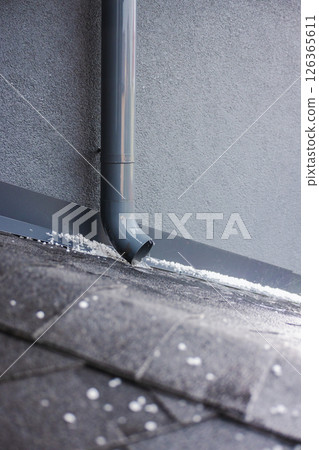 Close-up of water flowing from a gray downspout on a shingled roof covered with small hailstones. 126365611