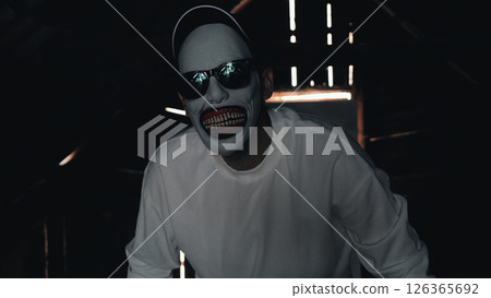 A man in a scary mask wearing a white sweatshirt and black cap is in a dimly lit attic with rays of light coming through the cracks. 126365692