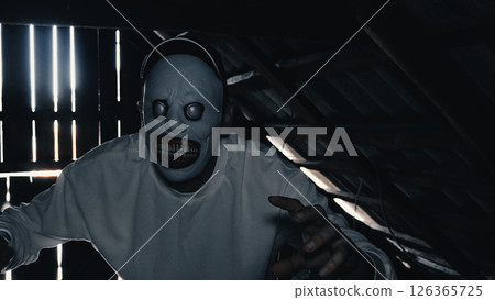 A creepy mask in a dark attic. A man wearing a terrifying mask with large empty eyes and a wide toothy grin. The man stands in a dark, attic room, creating a sense of unease. 126365725