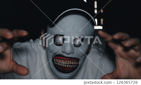 A man in a white hoodie and cap with a creepy mask on his face holds out both hands in a dark room, light streaming in through small holes in the background. 126365726