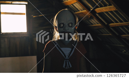 A young girl wearing a creepy mask with big eyes and a wide smile stands in a dark, shadow-covered attic. Her red sailor dress gives the scene a touch of disturbing innocence. 126365727