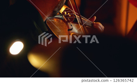 Close-up of a violin being played on the dimly lit Philharmonic stage. The background is blurred and has an orange colour from lit candles. Close-up of a violin being played on the dimly lit Philharmonic stage. The background is blurred and has an orange colour from lit candles. 126365742