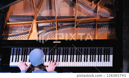 Top-down view of grand piano featuring copper-colored string frame and tuning pins above keyboard, with musician in blue jacket playing piano, hands spread across multiple octaves. 126365743