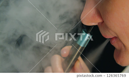 Close-up of a woman's face exhaling a cloud of vapor from an electronic cigarette. A hand holding a vape device, with thick clouds of vapor around it. 126365769